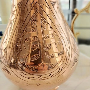 May include: A copper-colored pitcher with intricate engraved patterns. The design includes geometric shapes and a central stylized letter. The pitcher has a curved body, a handle, and a spout, showcasing a vintage aesthetic.