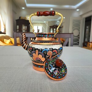 May include: A copper teapot with a floral design and a matching lid. The teapot has a gold handle and a wooden knob on the lid. The teapot is painted with black, white, red, blue, green, and orange colors.