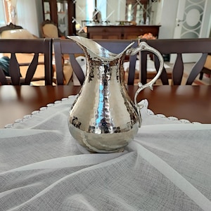 Puede incluir: Una jarra de metal martillado plateada con un asa decorativa. La jarra está sobre un mantel blanco.