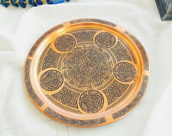 Puede incluir: Una bandeja redonda de cobre con intrincados diseños grabados en negro. La bandeja presenta un motivo floral central rodeado de diseños circulares más pequeños.
