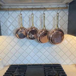 Puede incluir: Cinco ollas de color cobre cuelgan de un estante de latón sobre una estufa de gas negra. Las ollas tienen superficies martilladas y están suspendidas de ganchos en el estante.