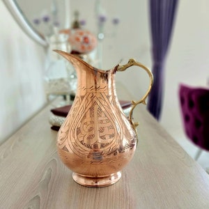 May include: A copper-colored pitcher with an ornate design etched into its surface. The pitcher has a long spout and a decorative handle. The pitcher is sitting on a light-colored wooden surface.