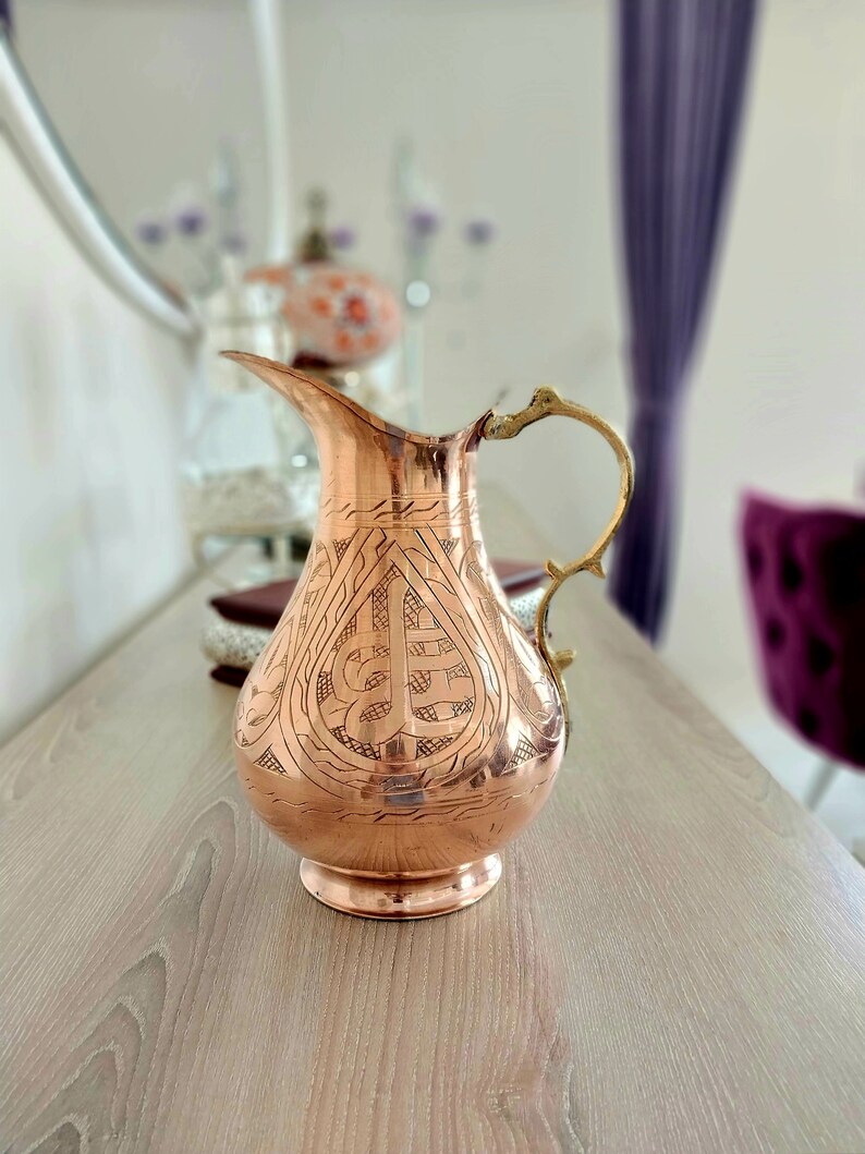 May include: A copper-colored pitcher with an ornate design etched into its surface. The pitcher has a long spout and a decorative handle. The pitcher is sitting on a light-colored wooden surface.