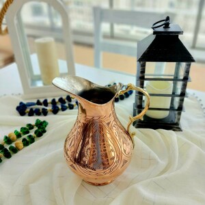 May include: A copper-colored pitcher with an ornate, etched design. The pitcher has a silver-colored spout and a gold-colored handle. The pitcher is sitting on a white tablecloth. A black lantern and a white candle holder are in the background.