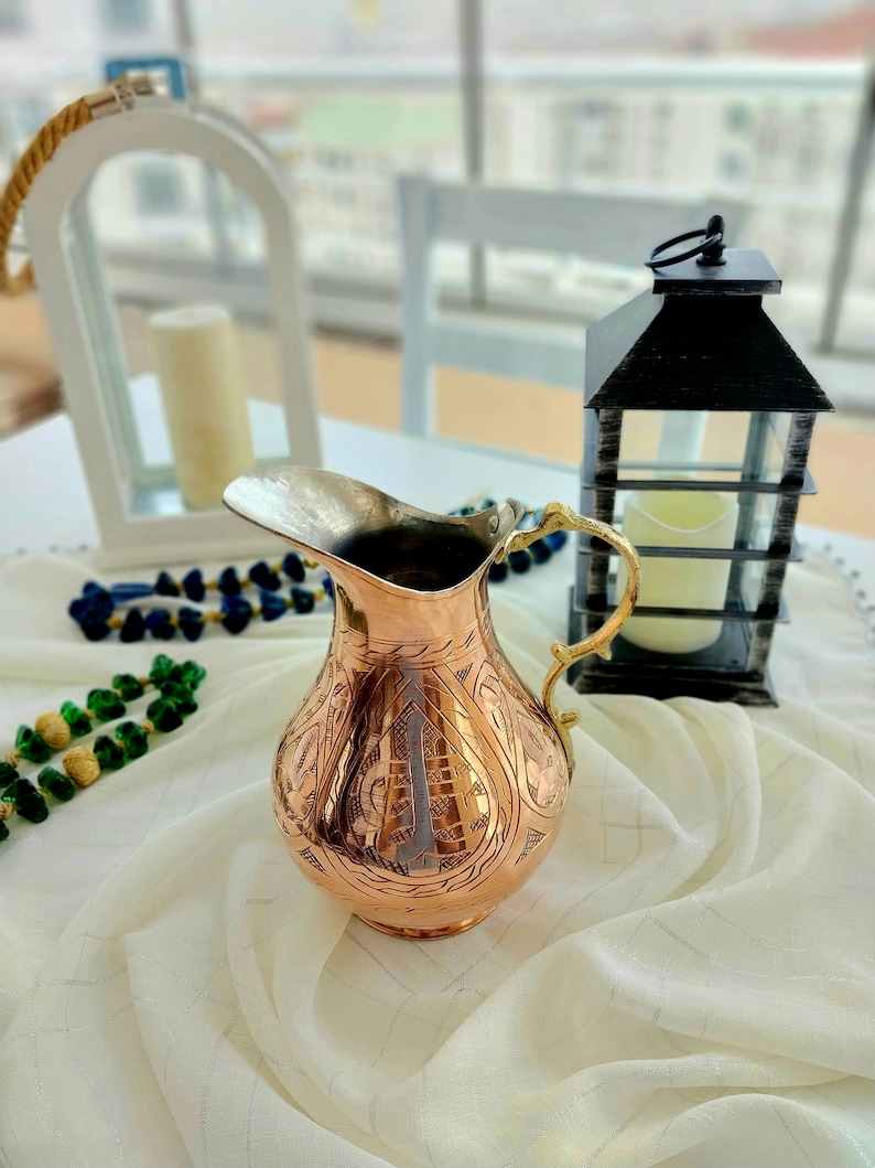 May include: A copper-colored pitcher with an ornate, etched design. The pitcher has a silver-colored spout and a gold-colored handle. The pitcher is sitting on a white tablecloth. A black lantern and a white candle holder are in the background.