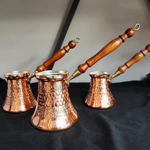 Könnte beinhalten: Drei Kaffeekannen aus Kupfer mit Holzgriffen. Die Kannen haben eine gehämmerte Oberfläche und aufwendige Verzierungen.