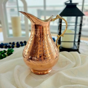 May include: A copper-colored pitcher with an ornate, etched design. The pitcher has a long spout and a decorative gold-colored handle. The background includes a white lantern, a black lantern, and a white cloth.