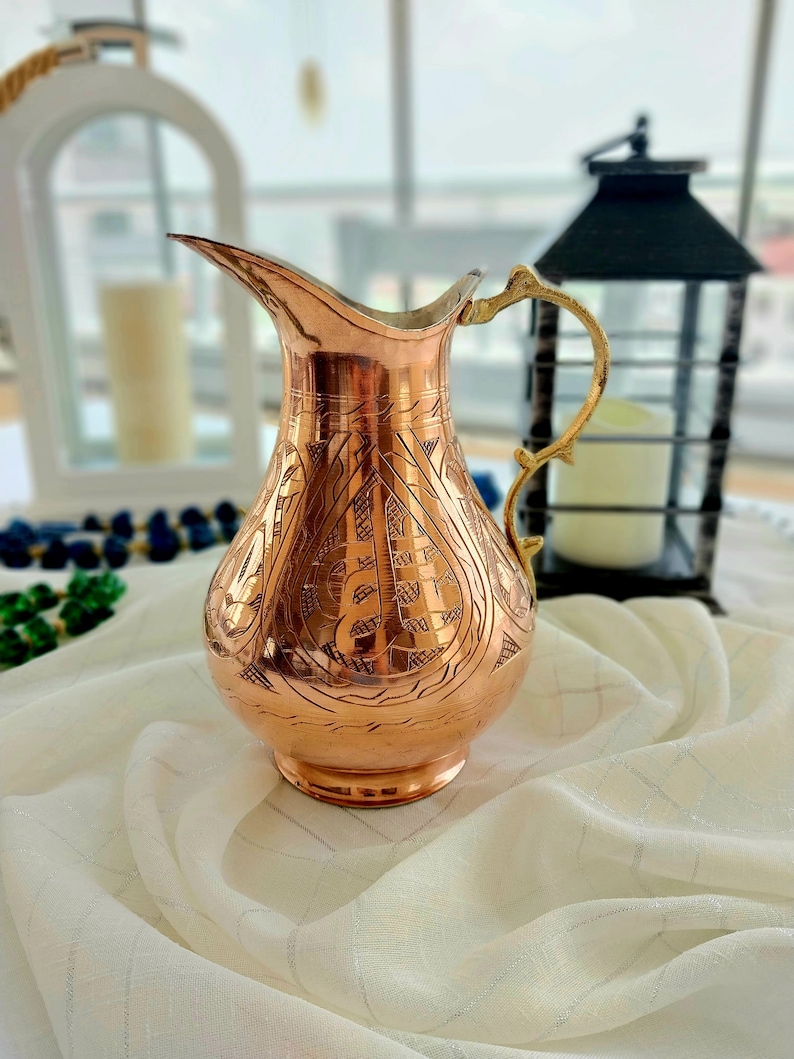 May include: A copper-colored pitcher with an ornate, etched design. The pitcher has a long spout and a decorative gold-colored handle. The background includes a white lantern, a black lantern, and a white cloth.