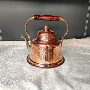 May include: A copper teapot with a hammered finish and a wooden handle. The teapot has a spout shaped like a swan's neck and a lid with a knob.