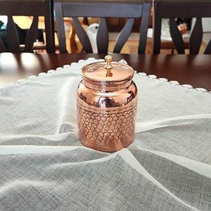 May include: A copper jar with a brass lid and a hammered texture. The jar is sitting on a white lace tablecloth.