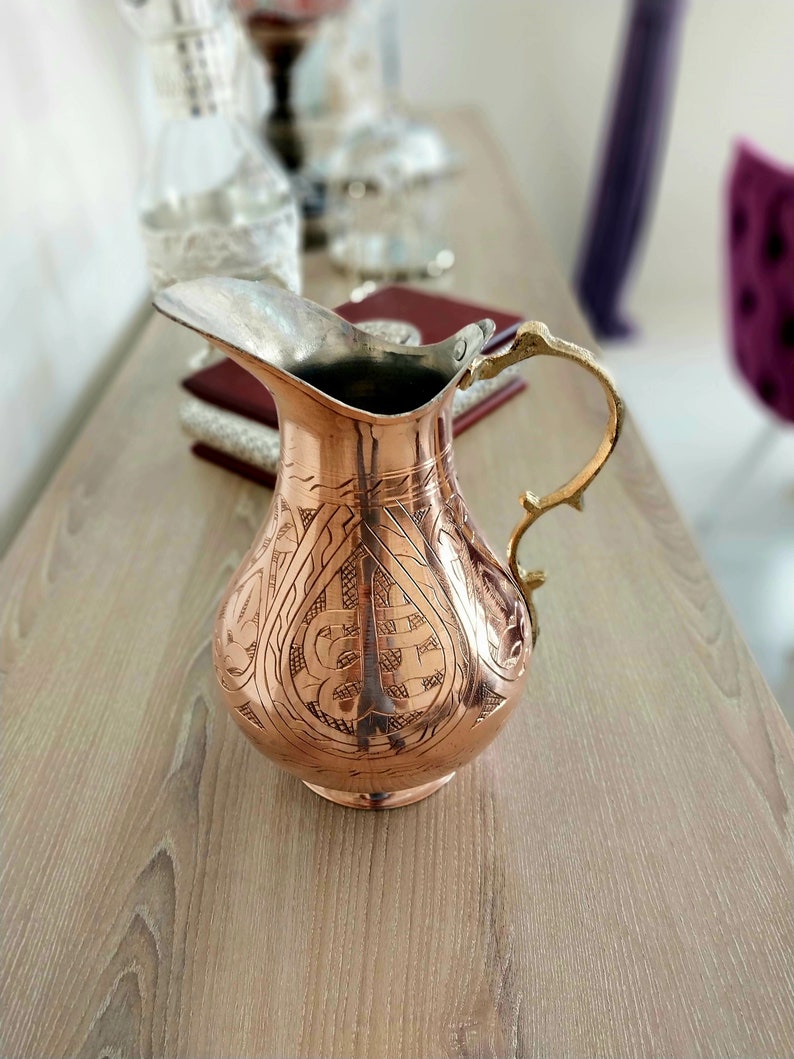 May include: A copper-colored pitcher with an ornate, engraved design. The pitcher has a silver-colored spout and a gold-colored handle. The pitcher is sitting on a wooden surface.