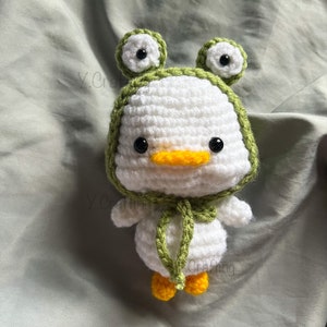 May include: White crocheted stuffed animal with a green frog hat. The frog hat has two large black eyes and a green brim. The stuffed animal has a yellow beak and black eyes.