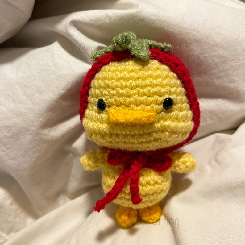May include: A crocheted yellow chick wearing a red and green crocheted strawberry hat. The chick has black eyes and a yellow beak.