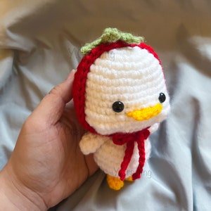 May include: A white crocheted stuffed animal wearing a red and green strawberry hat. The animal has a yellow beak and black eyes.