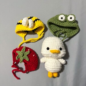 May include: Four crocheted hats in different designs: a bee, a frog, a strawberry, and a duck. The hats are all white with different colored accents.