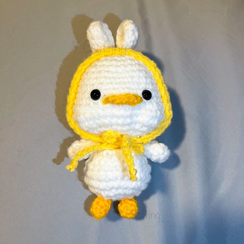 May include: A white crocheted stuffed animal with yellow accents. The animal has a duck-like beak and bunny ears.