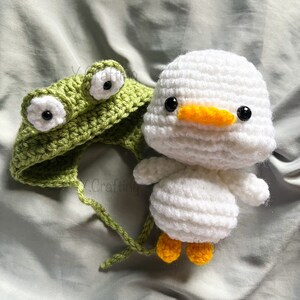 May include: A green crocheted frog hat with white eyes and a white crocheted duck with a yellow beak and black eyes.