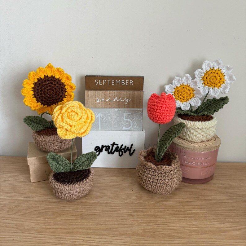 May include: Three crocheted flower arrangements in pots. The flowers are a sunflower, a rose, and a daisy. The pots are brown and beige. The background includes a wooden calendar with the date September 15th.