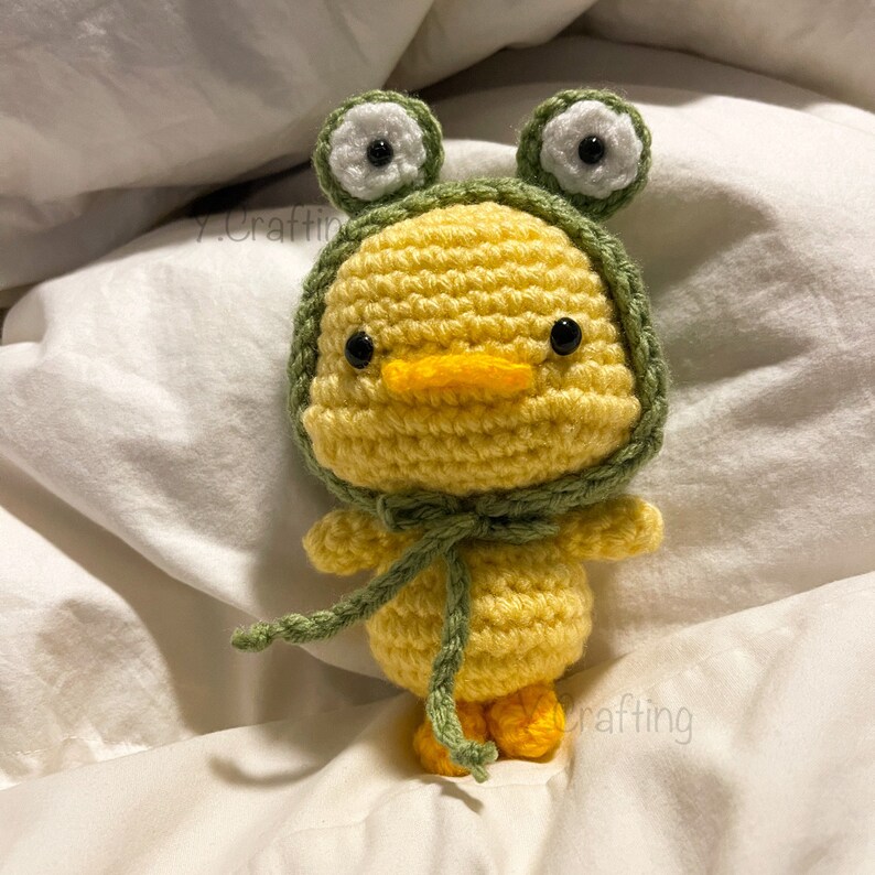 May include: A yellow crocheted duck wearing a green crocheted frog hat with white eyes. The duck has a yellow beak and black eyes.