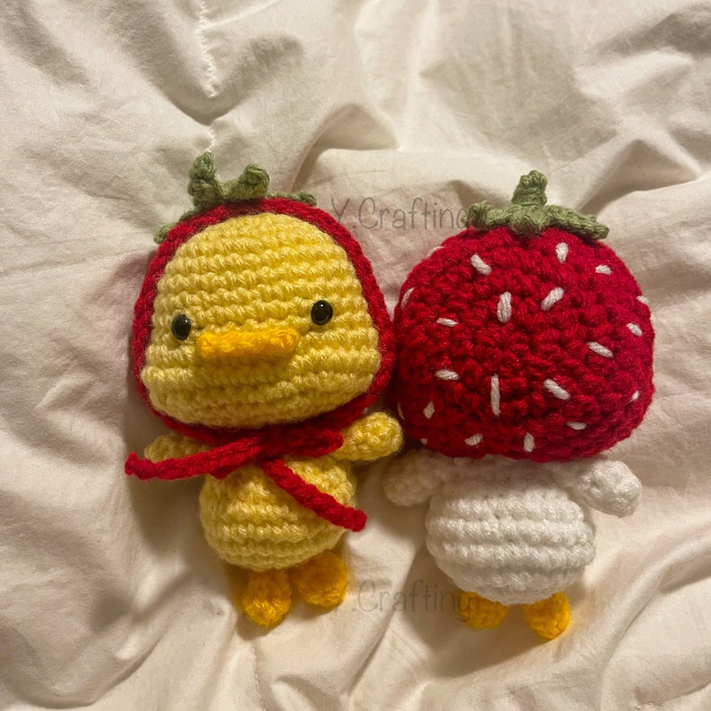 May include: Two crocheted stuffed toys, one is a yellow chick wearing a red strawberry hat and the other is a white strawberry with a red top.