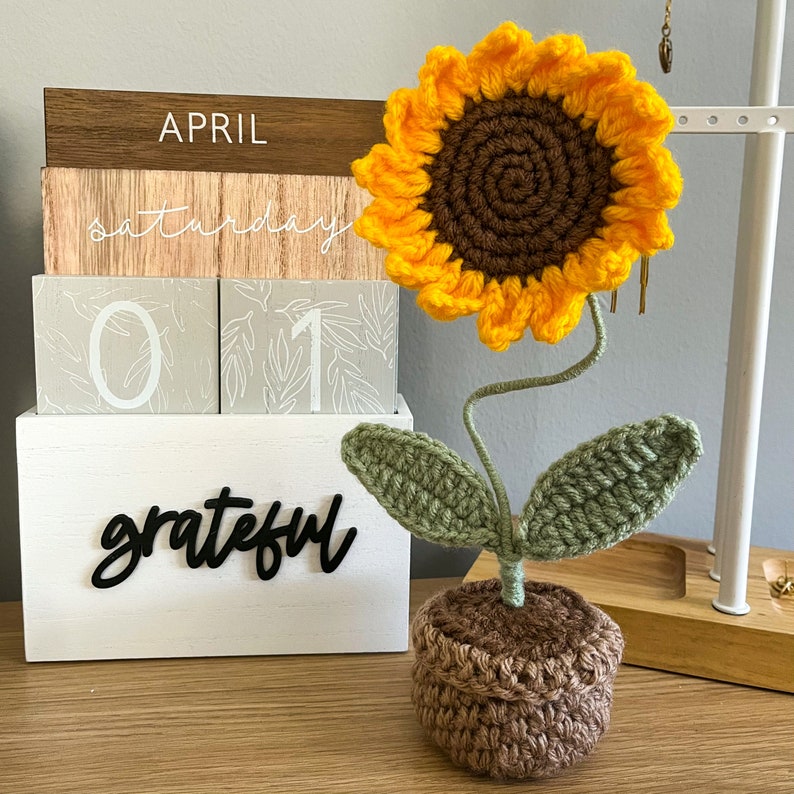 May include: A crocheted sunflower with green leaves and a brown pot. The sunflower is on a wooden stand with a white box that says "grateful" and a wooden block calendar that says "April Saturday 01".
