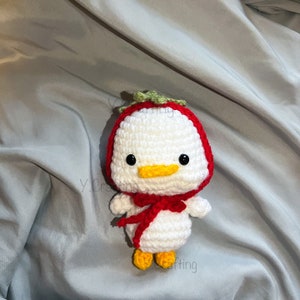 May include: A white crocheted duck wearing a red and green strawberry costume. The duck has black eyes, a yellow beak, and yellow feet.
