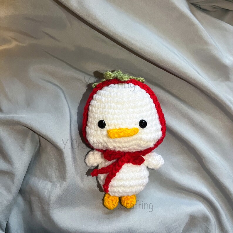 May include: A white crocheted duck wearing a red and green strawberry costume. The duck has black eyes, a yellow beak, and yellow feet.
