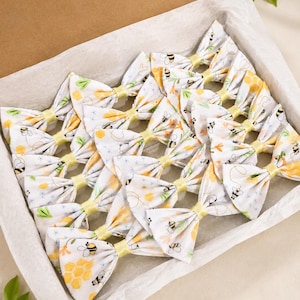 May include: A collection of fabric bows arranged in a cardboard box lined with white tissue paper. Each bow features a white background with a repeating pattern of yellow honeycombs, small black and yellow bees, and light blue star accents.