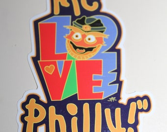 May include: A sticker with the text "Me Love Philly!" in a colorful font. The letter "O" in "Love" is replaced with a cartoon character with orange fur, a red nose, and a blue hat. The character has a big smile and is wearing a red and green shirt.