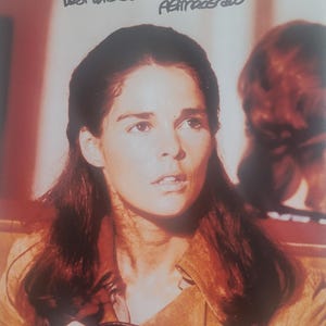 以下が含まれることがあります： 茶色のスエードジャケットを着た、長い茶色の髪の女性をクローズアップで写した写真。彼女は黒い革のストラップを手に持っている。写真は「ベストウィッシュ＆ピース、アリ・マッグロー」とサインされている。