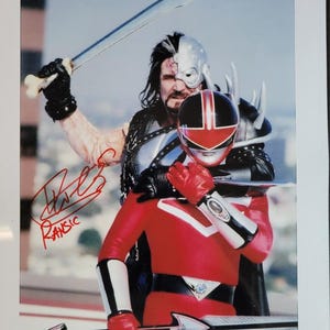 May include: A signed photograph featuring two costumed characters. One character wears a red and black helmet and suit, holding a sword. The other character, with a half-mask, holds a sword and bone. The signature reads "Ransic".