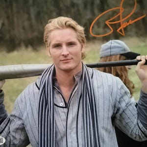 May include: A photograph featuring a blonde individual wearing a grey pinstriped button-down shirt and a matching striped scarf, holding a metallic baseball bat across their shoulders. An orange signature is visible in the upper right corner.