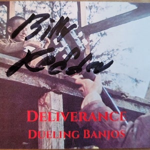 May include: A black and white photo of a scene from the movie Deliverance. The text "Deliverance Dueling Banjos" is printed in red at the bottom of the photo. The photo is signed by actor,  Bill  Redden.