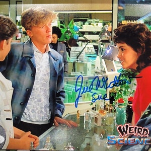 May include: A signed photograph of the cast from the film "Weird Science". The image features three people, two men and one woman, standing in a shop. The woman is wearing a pink shirt and the man on the left is wearing a white shirt with a blue and white pattern. The man on the right is wearing a blue jacket over a white shirt with a blue and white pattern. The photograph is signed by the actress, "Irid Wicker Sue".