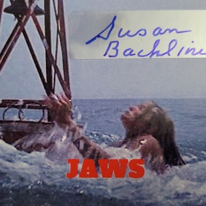 Puede incluir: Una fotografía firmada de Susan Backlinie, la actriz que interpretó a la primera víctima en la película Tiburón. La imagen la muestra en el agua, con la palabra "JAWS" en letras rojas debajo de ella.