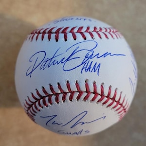 May include: A white baseball with red stitching and multiple blue signatures. The ball has a smooth surface and is likely a collectible item. The signatures appear to be from baseball players.