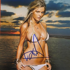 Könnte beinhalten: Eine Frau in einem weißen Bikini mit goldenen Akzenten posiert bei Sonnenuntergang am Strand. Sie hat blonde Haare und schaut in die Kamera. Das Bild ist mit einer blauen Unterschrift versehen.