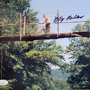 May include: A black and white photo of a man walking across a wooden bridge in a forest. The man is carrying a bucket and is wearing a light-colored shirt and pants. The bridge is made of logs and rope and is suspended over a deep ravine. The signature "Billy Keller" is written in black ink in the upper right corner of the photo.
