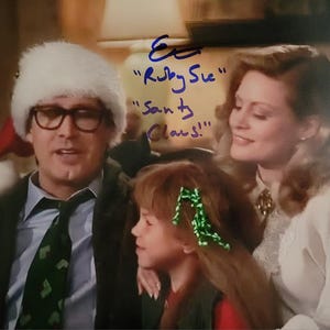 Puede incluir: Retrato familiar con un hombre con gorro de Papá Noel y gafas, una mujer y un niño con accesorios para el pelo verdes. El hombre lleva traje y corbata. La imagen tiene texto escrito a mano que dice "Ruby Sue" y "Santa Claus!".