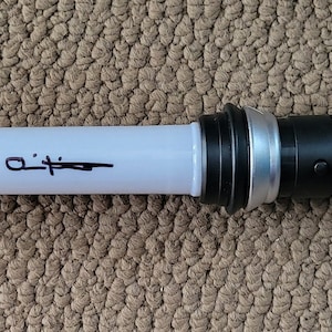 May include: A lightsaber prop with a white blade and a black and silver handle. The blade has a signature in black ink. The handle features a black button and a silver ribbed design. The lightsaber is lying on a brown surface.