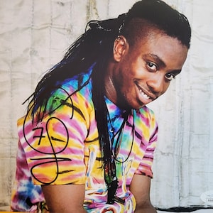 May include: A black and white photograph of a man with long dreadlocks wearing a tie-dye shirt and smiling. The man is signing his name on the photo.