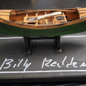 May include: A small wooden model of a green and brown canoe on a black wooden base with the name "Billy Reiden" written in white paint.