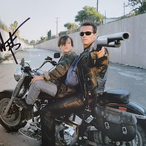 May include: A photograph of two people on a black motorcycle. One person is wearing a black leather jacket and sunglasses, holding a shotgun. The other person is wearing a camouflage jacket and jeans. The image is signed in the upper left corner.