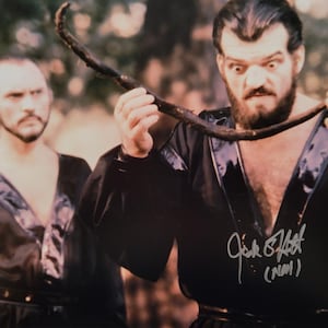 May include: A glossy photograph featuring two individuals in black robes with shiny lapels. The foreground figure has a dark beard and wide eyes, holding a long, dark, curved object. A white signature is visible on the lower right side of the image.
