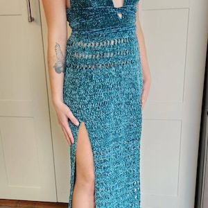 May include: A teal blue crochet maxi dress with a halter neckline and a thigh-high slit. The dress is made of a soft, fuzzy yarn and has a crocheted pattern throughout.