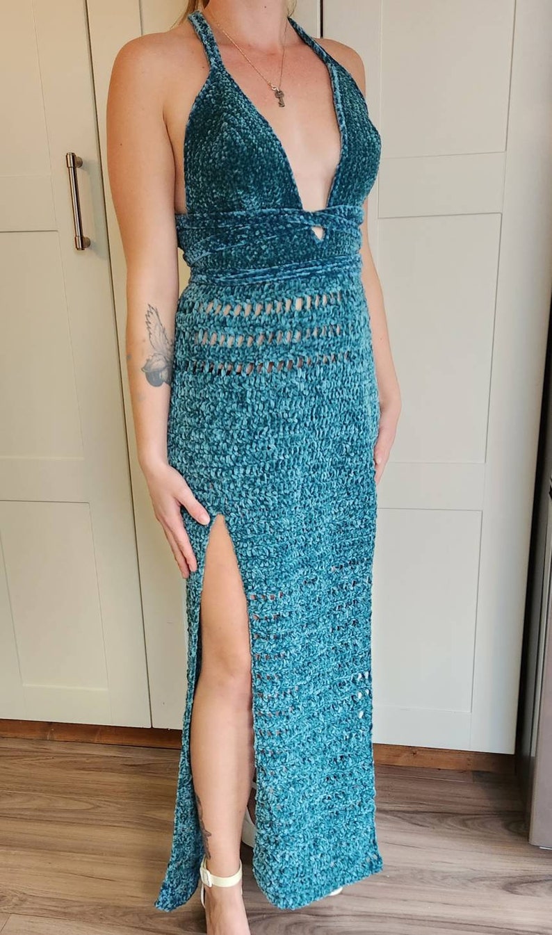 May include: A teal blue crochet maxi dress with a halter neckline and a thigh-high slit. The dress is made of a soft, fuzzy yarn and has a crocheted pattern throughout.