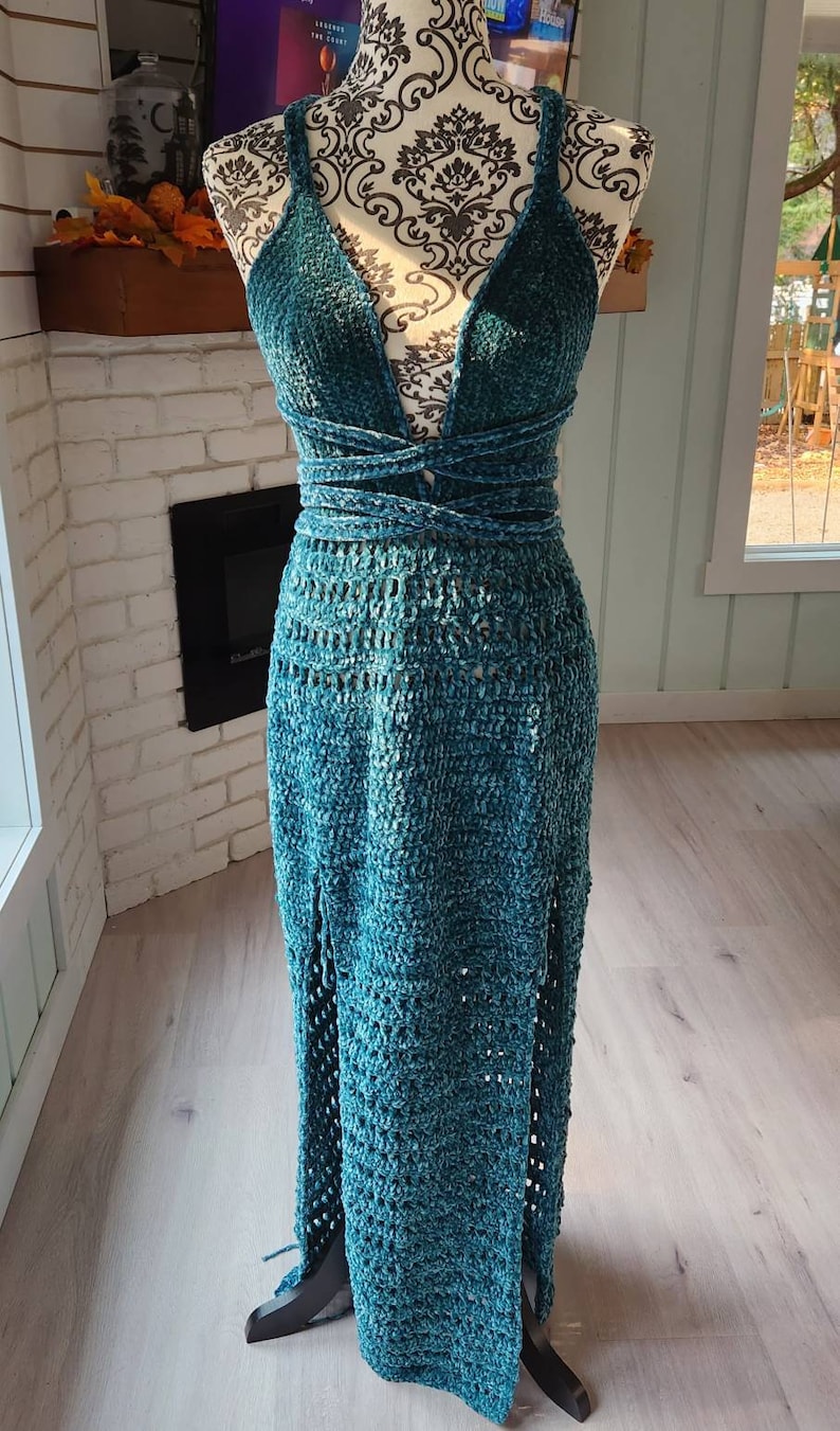 May include: A teal blue crochet maxi dress with a deep V-neckline and a wrap-style bodice. The dress has a crocheted skirt with a slit on the front.