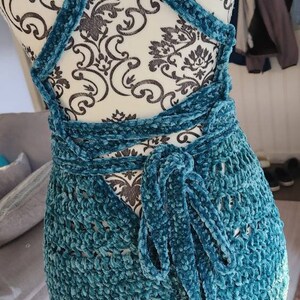 May include: A teal blue crocheted dress with a velvet texture. The dress has a criss-cross strap design in the back and ties at the waist.