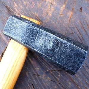 May include: A heavy-duty hammer with a dark, textured metal head and a light-colored wooden handle. The hammer head has a hexagonal shape and appears to be made of forged steel. The handle is smooth and tapered, providing a comfortable grip for various construction tasks.