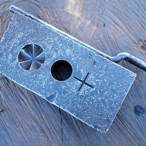 May include: A weathered, rectangular metal object with a handle. The object has a starburst-shaped hole, a circular hole, and a cross-shaped marking. The metal has a textured, aged appearance, suggesting it is a vintage or handmade item.